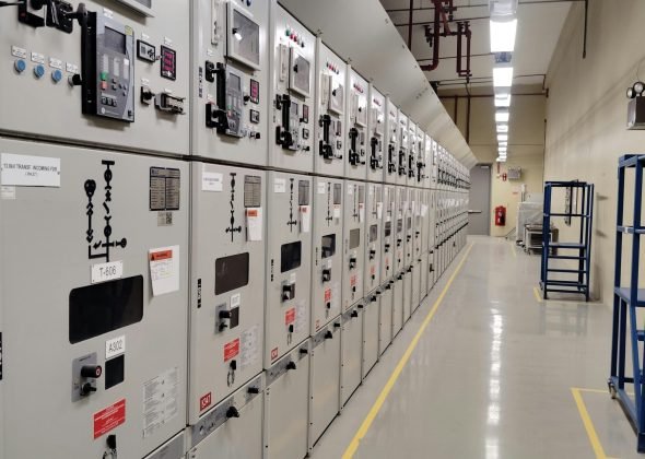 Long row of control panels in an industrial electrical control room, well-lit and spacious.